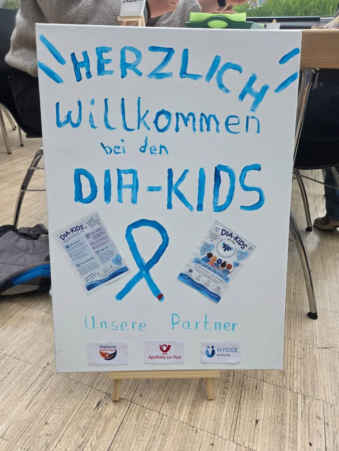 Handgemaltes Schild „Herzlich Willkommen bei den DIA-KIDS“ mit blauer Schleife, Flyern und den Logos der Partner